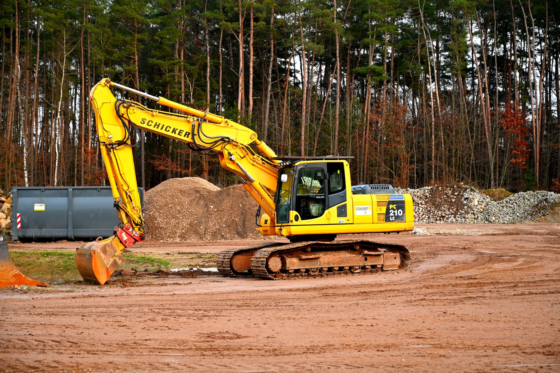A yellow tracked excavator standing on churned bare ground at a cleared site, with heaped spoil and graded stone behind it and pines along the back of the plot.