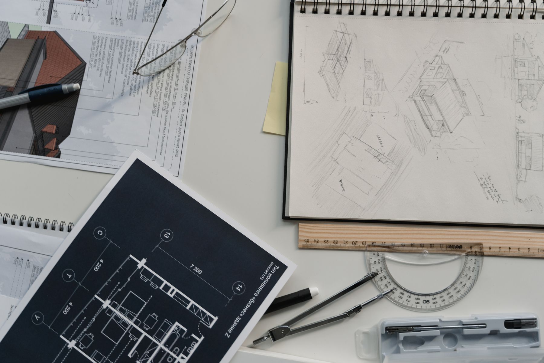 A designer’s desk seen from above with a sketchbook of hand-drawn perspective studies, a blue printed floor plan, a set square, protractor, dividers and pencils.
