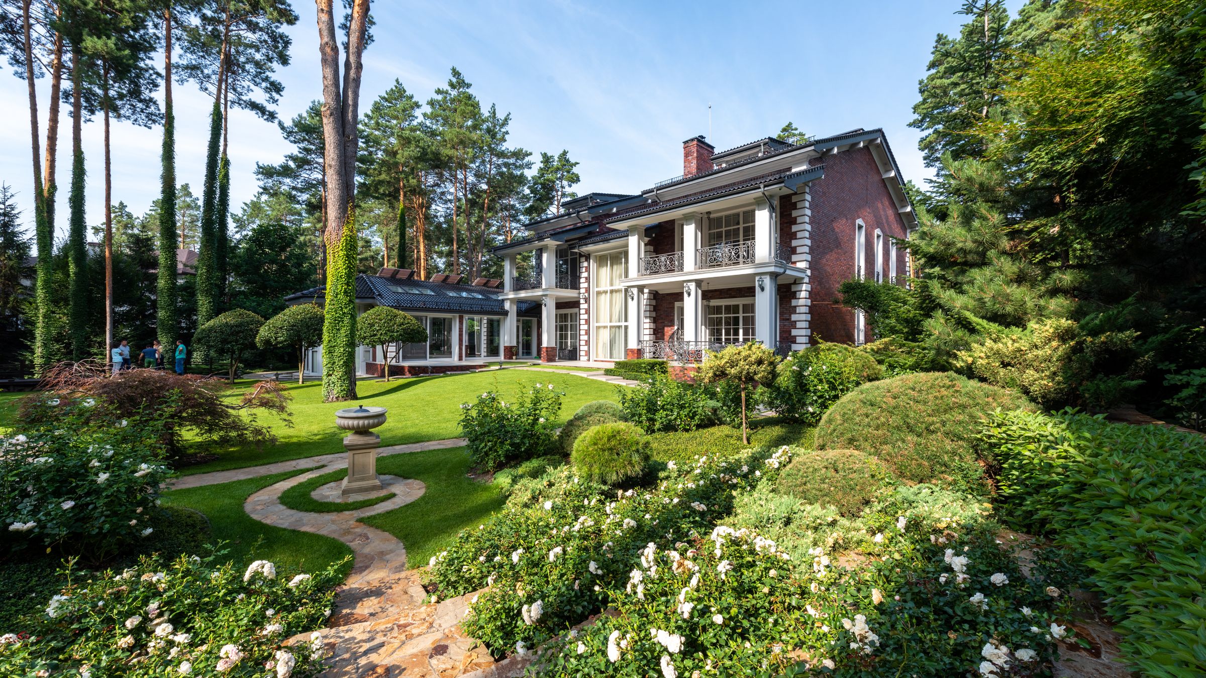 A mature private garden with clipped topiary, massed white roses and a stone path curving past a small stone fountain towards a large house.