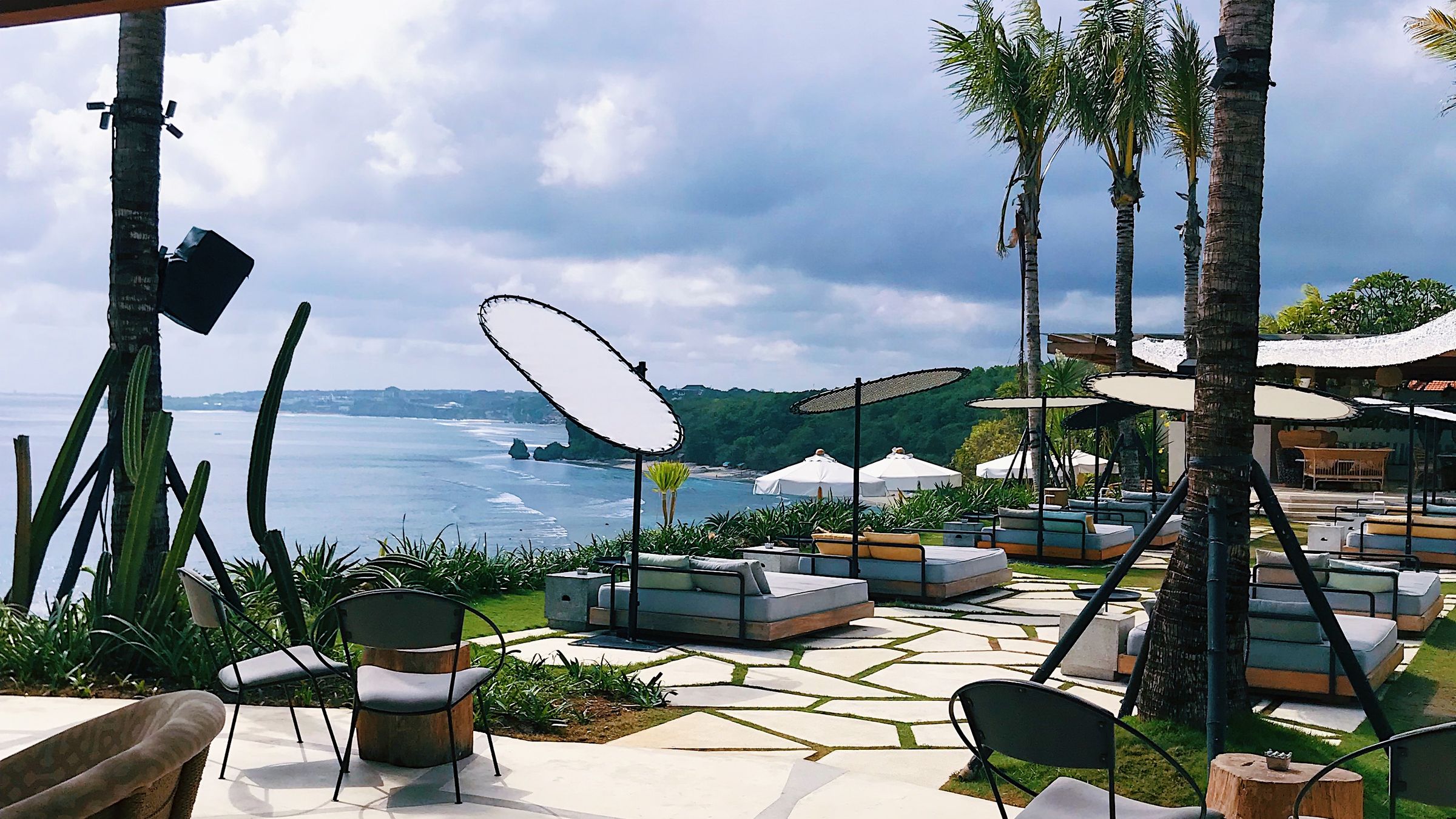 A resort terrace above the sea with day beds and parasols under palms on flagstone paving.