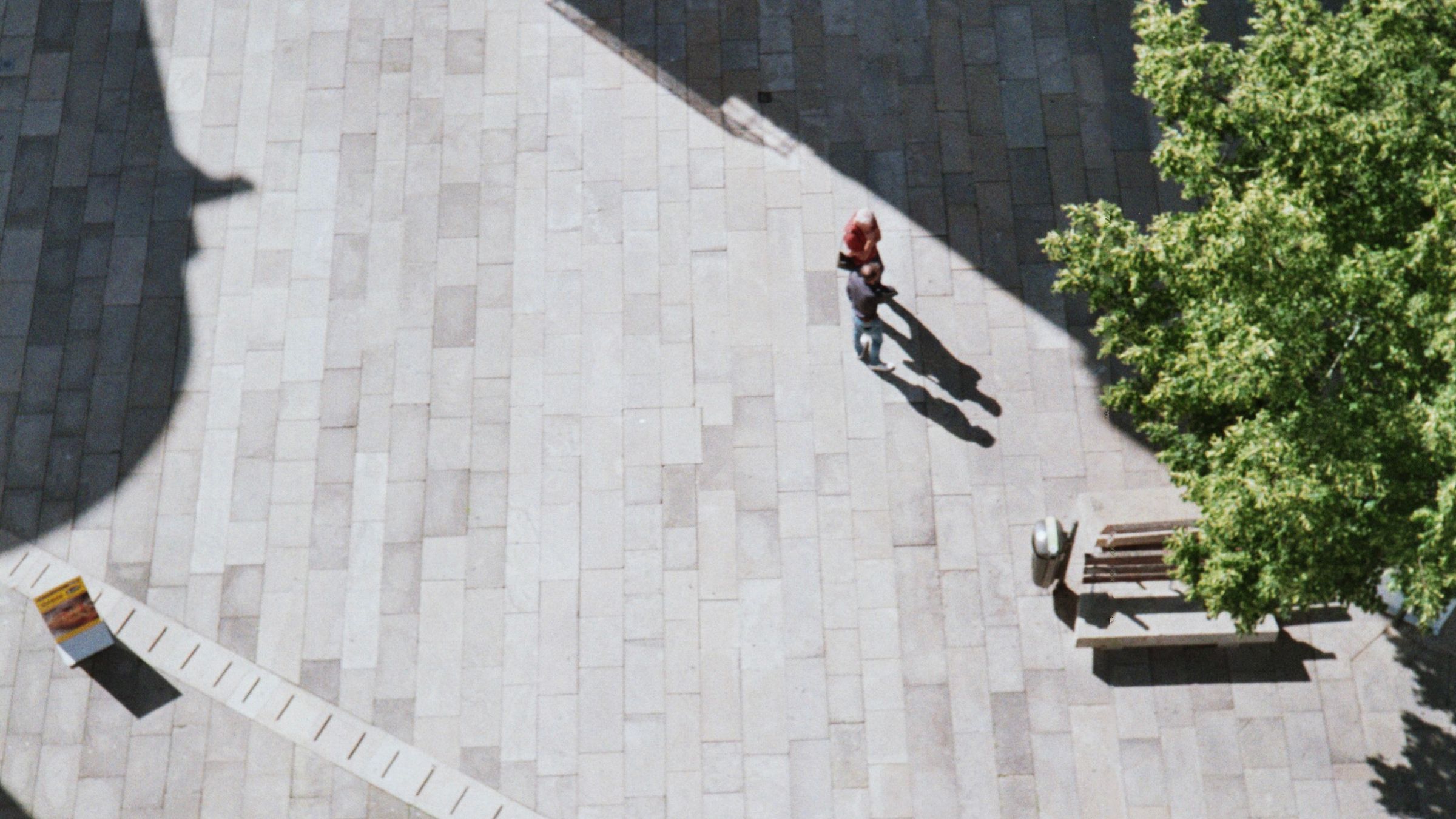 A large paved civic square seen from above with one figure walking across it, and a line of trees and benches down one side.