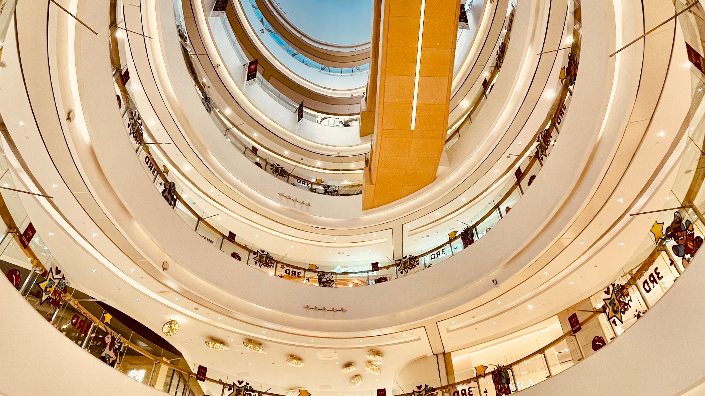 A shopping centre atrium under a circular skylight, with balconies stepping back on each level and escalators crossing the void.